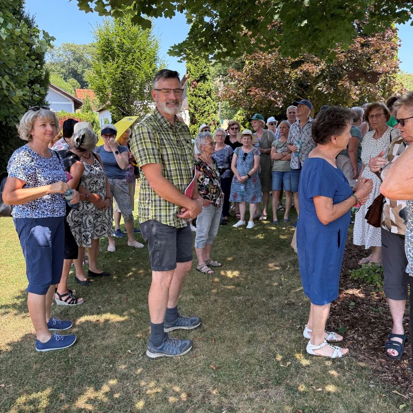 Eine Gruppe von Menschen steht in einem Garten unter einem Baum und trägt lässige Sommerkleidung. Einige haben Hüte und Brillen auf, und einige tragen Handtaschen. Die Atmosphäre scheint entspannt und freundlich.