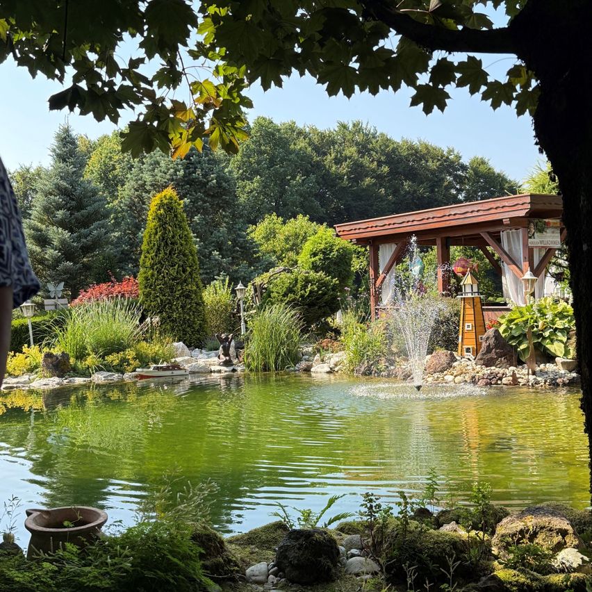 Eine friedliche Gartenszene mit einem Teich und einem Springbrunnen. Der Teich ist von üppiger Grünfläche und Felsen umgeben. Ein hölzerner Pavillon steht in der Nähe des Teiches. Bäume und Sträucher tragen zur friedlichen Atmosphäre bei.