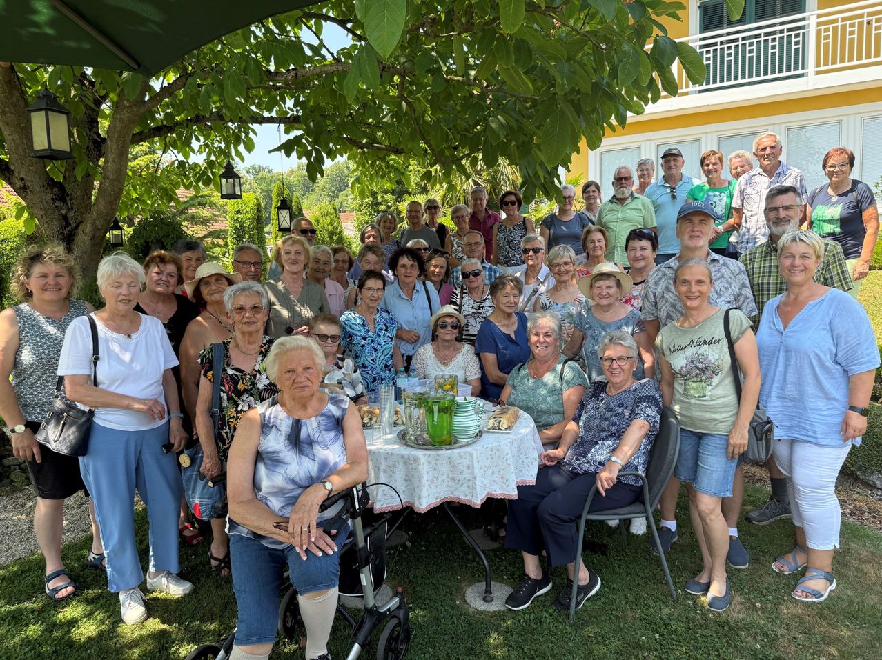 Eine große Gruppe älterer Menschen versammelt sich für ein Foto unter einem Baum im Garten, mit einem Tisch voller Speisen und Getränken davor.
