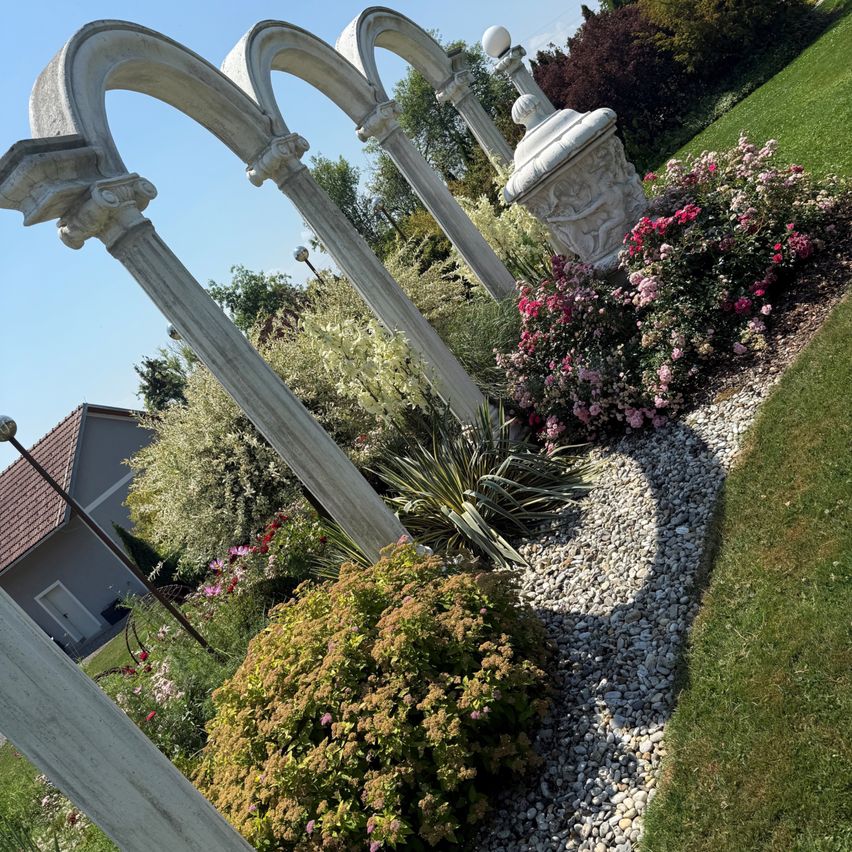 Ein Garten mit rosa und weißen Blumen und Betonbögen im Hintergrund. Es gibt Büsche, Kies und ein Haus mit einem braunen Dach in der Ferne.