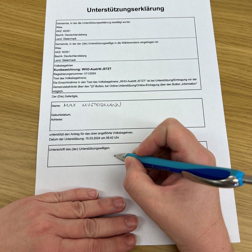 Eine Hand hält einen Stift und schreibt auf einem weißen Blatt Papier mit der Überschrift 'Unterstützungserklärung'. Das Formular enthält Felder für Name, Geburtsdatum, Adresse und eine Unterschrift. Darunter befinden sich Angaben zu einer Gemeinde, Registrierungsnummer und einem Datum. Das Papier liegt auf einer Holzoberfläche.