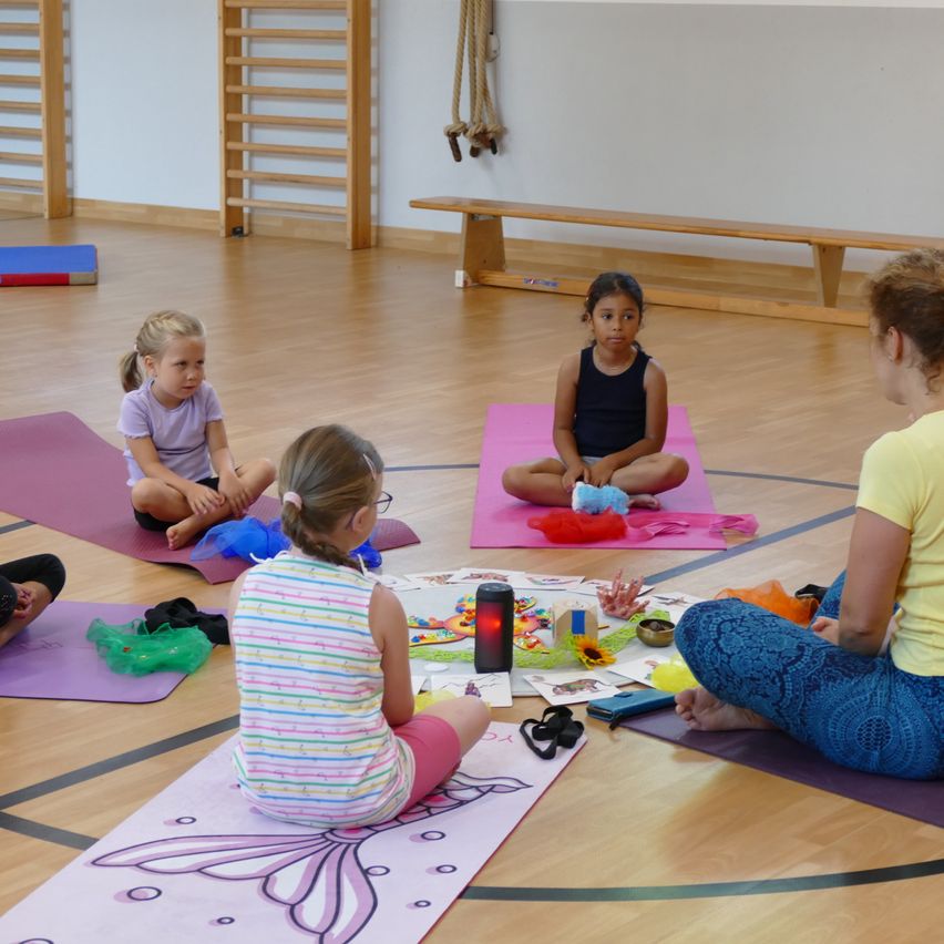Drei Mädchen und eine Frau sitzen im Kreis auf Yogamatten. Die Frau schaut auf die Mädchen, mit Spielzeug und einer Laterne auf dem Boden zwischen ihnen. Ein Mädchen trägt eine Brille und Tüllröcke.