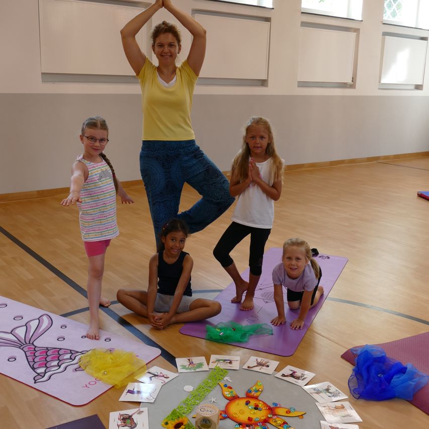 Eine Frau steht auf einem Bein in einer Yoga-Pose, während vier Mädchen, jedes auf einer Yogamatte, ihre Pose nachahmen. Der Raum hat Holzböden und weiße Wände mit Fenstern. Es gibt verschiedene Yoga-Zubehör und Matten auf dem Boden.