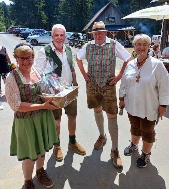 Vier Personen in traditioneller Kleidung posieren für ein Foto im Freien mit einem Geschenkkorb. Hinter ihnen sind Autos geparkt und ein Gebäude ist sichtbar.