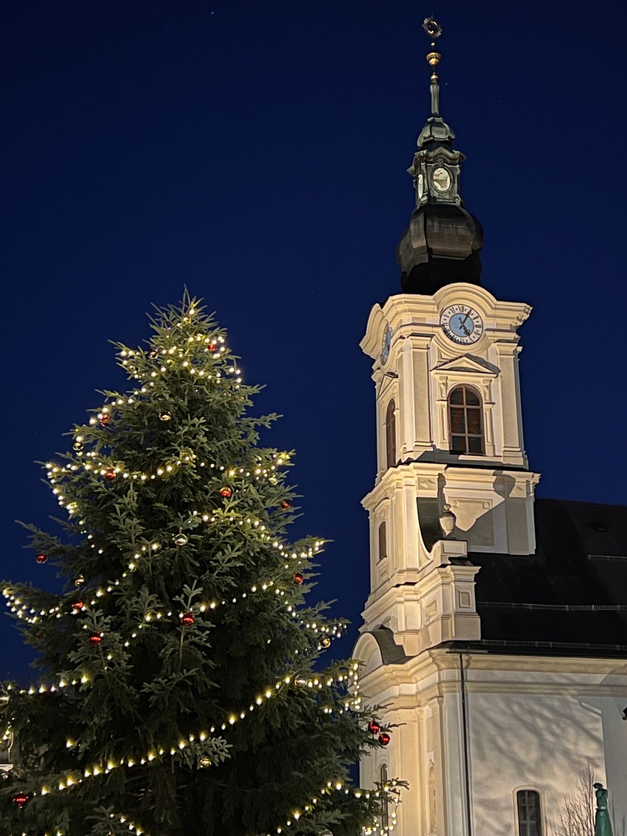 Ein Weihnachtsbaum mit roten Ornamenten und Lichtern steht vor einem hohen Kirchturm. Der Turm hat eine Uhr und ist nachts beleuchtet.