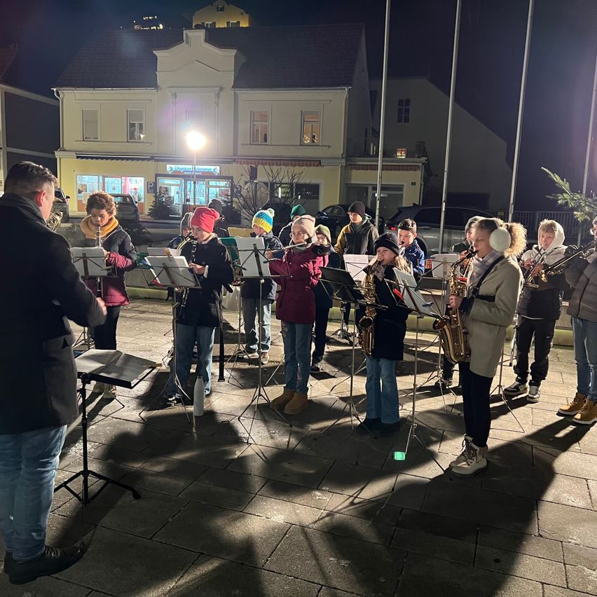 Eine Gruppe von Musikern steht nachts auf einem gepflasterten Bürgersteig und spielt Instrumente wie Saxophone und Trompeten unter einer Straßenlaterne. Im Hintergrund befindet sich ein Gebäude mit Fenstern und einem Schild.