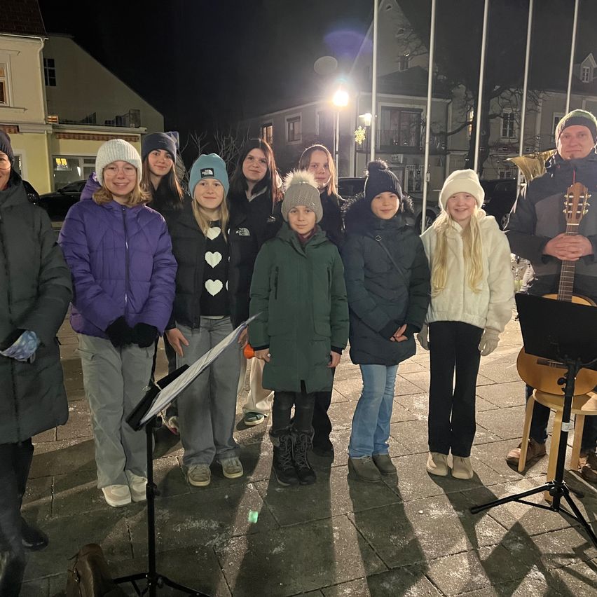 Eine Gruppe von Menschen steht in einer Reihe, möglicherweise singend, vor einem Gebäude bei Nacht. Ein Mann im Hintergrund spielt Gitarre.