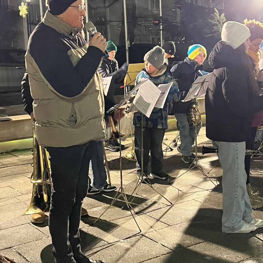 Eine Gruppe von Musikern, einschließlich eines Mannes mit Mikrofon, steht auf einem gepflasterten Bürgersteig und tritt bei Nacht auf. Sie sind warm gekleidet und halten Notenblätter.