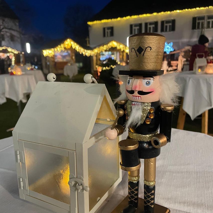 Ein dekorativer Nussknacker steht neben einer leuchtenden Laterne. Sie befinden sich in einer festlichen Außenumgebung mit Tischen, die mit weißen Tischdecken bedeckt sind, und einem Haus mit beleuchteten Fenstern.