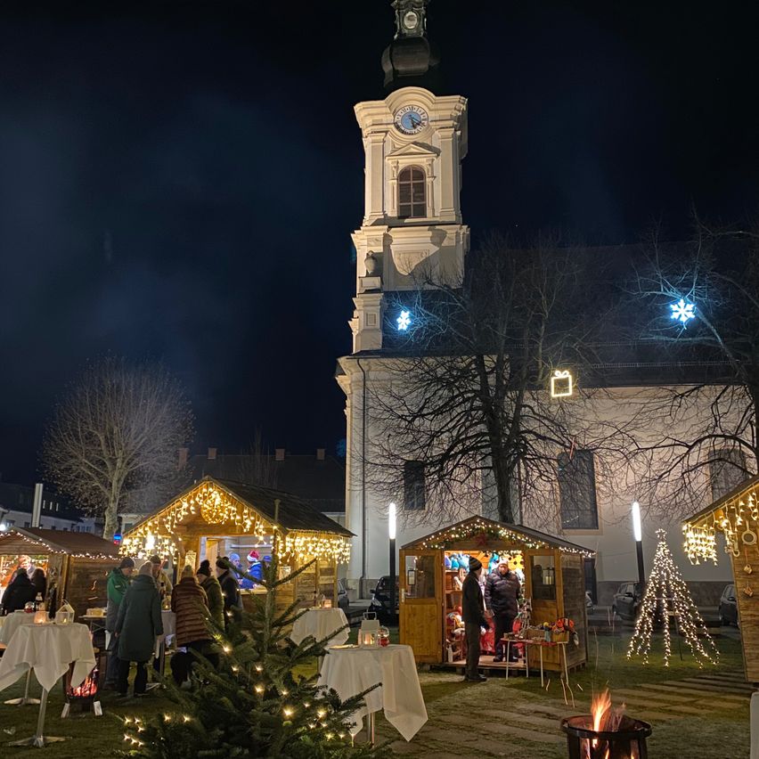 Ein Weihnachtsmarkt findet vor einer Kirche bei Nacht statt. Die Kirche ist mit Lichtern und einer Uhr geschmückt. Es sind mehrere Stände mit Tischen aufgestellt, einer mit einem Weihnachtsbaum und einem Feuerkorb.