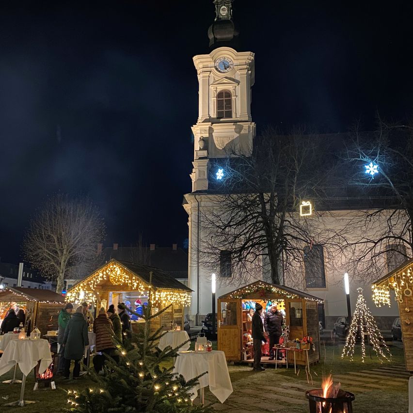 Eine Nachtszene eines Weihnachtsmarktes vor einer Kirche mit einem Glockenturm, geschmückt mit Lichtern und Bäumen, und Menschen, die um Stände herumstehen.