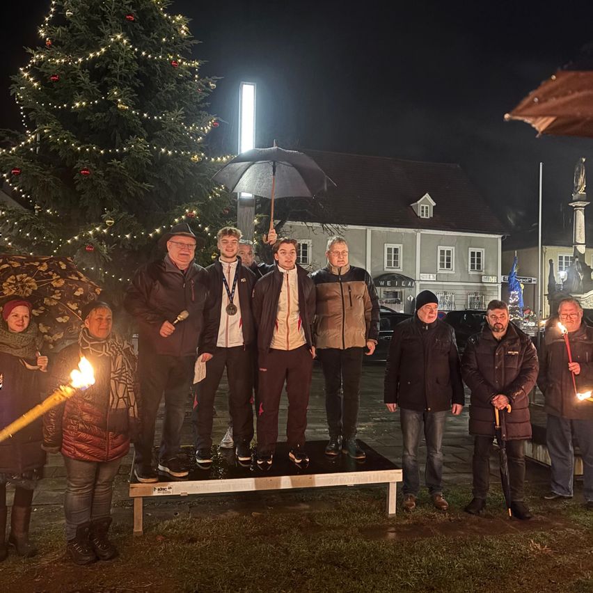 Eine Gruppe von Menschen steht bei Nacht auf einer Bühne mit einem Weihnachtsbaum im Hintergrund, hält Kerzen und Regenschirme. Ein Gebäude und eine Statue sind im Hintergrund.