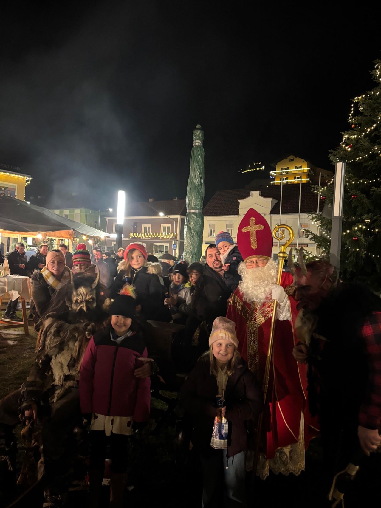 Eine fröhliche Menschenmenge versammelt sich nachts um den Weihnachtsmann, mit Gebäuden, einem Weihnachtsbaum und einer Person in einem Wolfskostüm im Hintergrund.