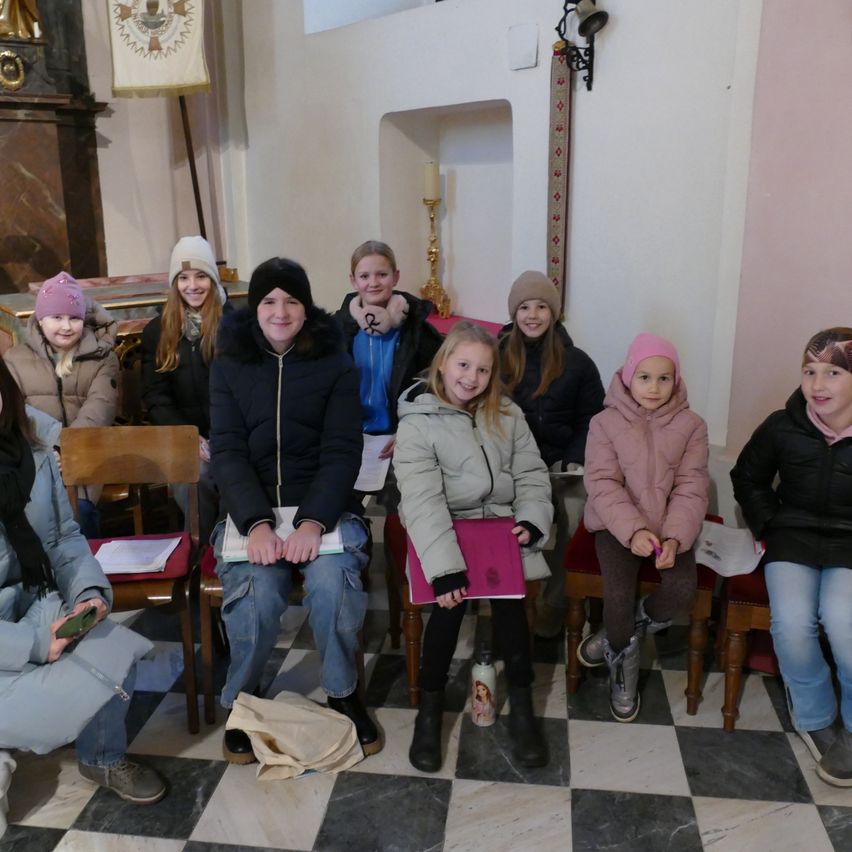 Eine Gruppe von Kindern und eine erwachsene Frau sitzen auf Stühlen in einer Kirche. Sie sind alle warm gekleidet. Der Raum hat einen gemusterten Boden und eine Kerze auf einem Kerzenständer.