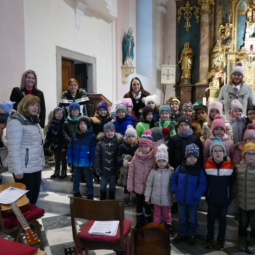 Eine Gruppe von Kindern in Winterkleidung posiert vor einem Kirchenaltar mit Erwachsenen, darunter eine Frau mit einer Gitarre. Die Kinder lächeln und einige tragen Mützen. Der Altar hat Statuen und ein goldenes Design.