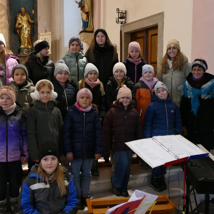 Eine Gruppe von Kindern und Erwachsenen steht in einer Kirche, alle tragen Winterkleidung und einige Mützen. Musiknoten und Bücher sind auf dem Boden zu sehen.