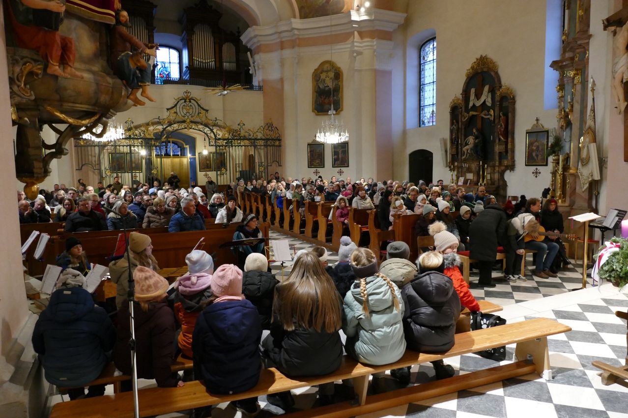 Eine Kirche voller Menschen, einige auf hölzernen Bänken sitzend, andere stehend. Ein Chor tritt vorne auf, mit Kronleuchtern an der Decke. Kunstwerke schmücken die Wände.