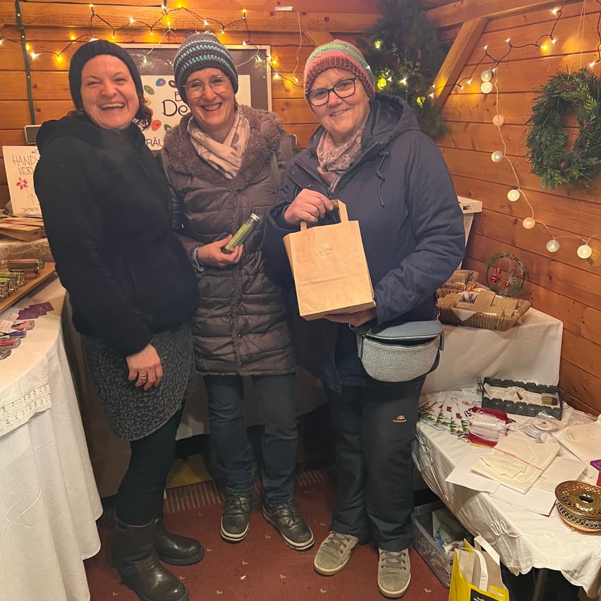 Drei Frauen stehen zusammen in einem festlichen Ambiente. Sie lächeln und scheinen für ein Foto zu posieren. Eine hält einen Papierbeutel, eine andere eine kleine Flasche und die dritte hat eine Handtasche. Hinter ihnen befinden sich zwei Tische mit verschiedenen Gegenständen, einschließlich einer Girlande. Der Hintergrund besteht aus Holzwänden und Lichterketten.