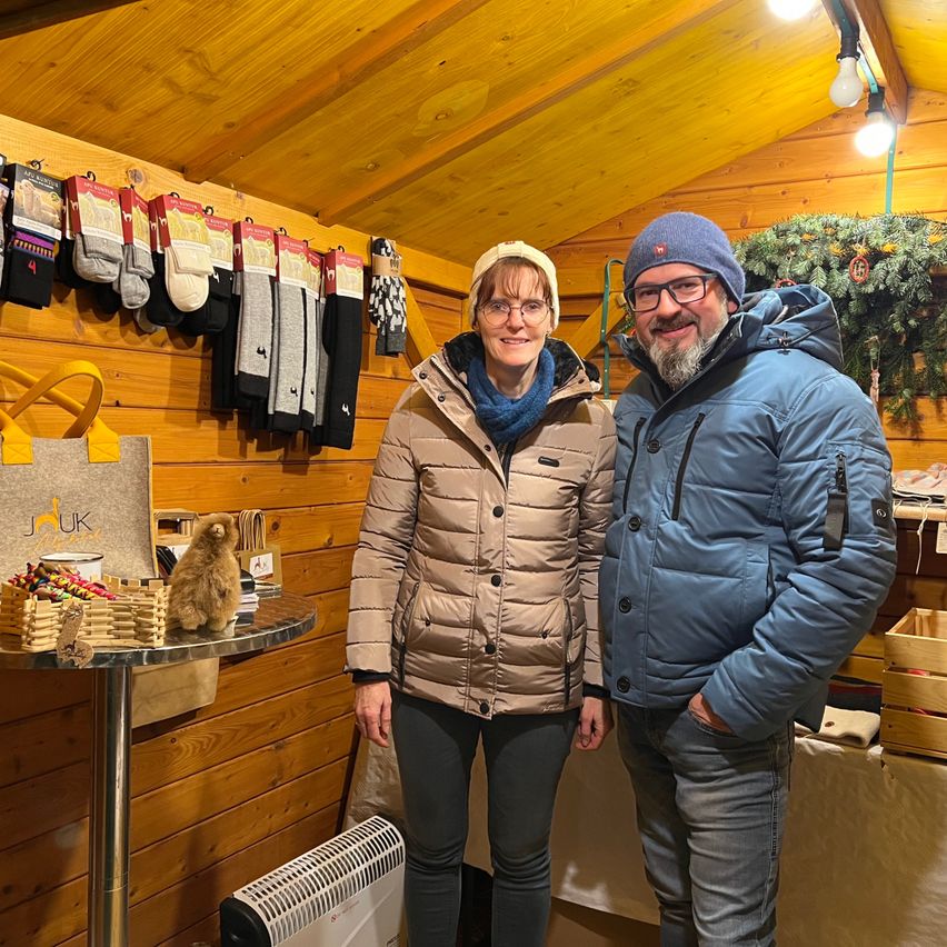 Ein Mann und eine Frau stehen in einer kleinen Holzhütte mit Winterkleidung und Socken an den Wänden, lächeln für ein Foto.