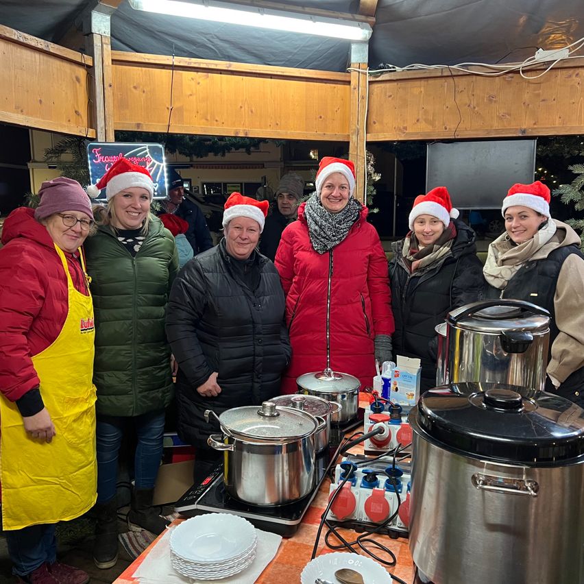 Eine Gruppe von Frauen in Weihnachtsmützen und Wintermänteln steht um einen Tisch mit Kochtöpfen und Tellern herum, lächelt und posiert für ein Foto in einem hölzernen Raum.