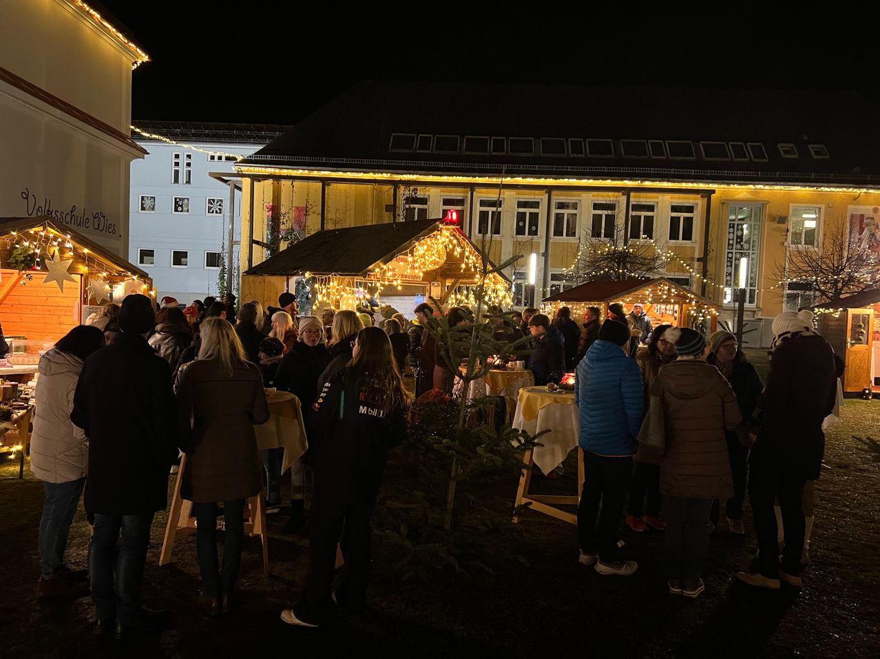 Eine nächtliche Szene auf einem Weihnachtsmarkt mit festlichen Lichtern und einer Menschenmenge, die sich um Stände und einen geschmückten Tannenbaum versammelt. Gebäude mit gelbem Licht und einer verschneiten Kulisse.