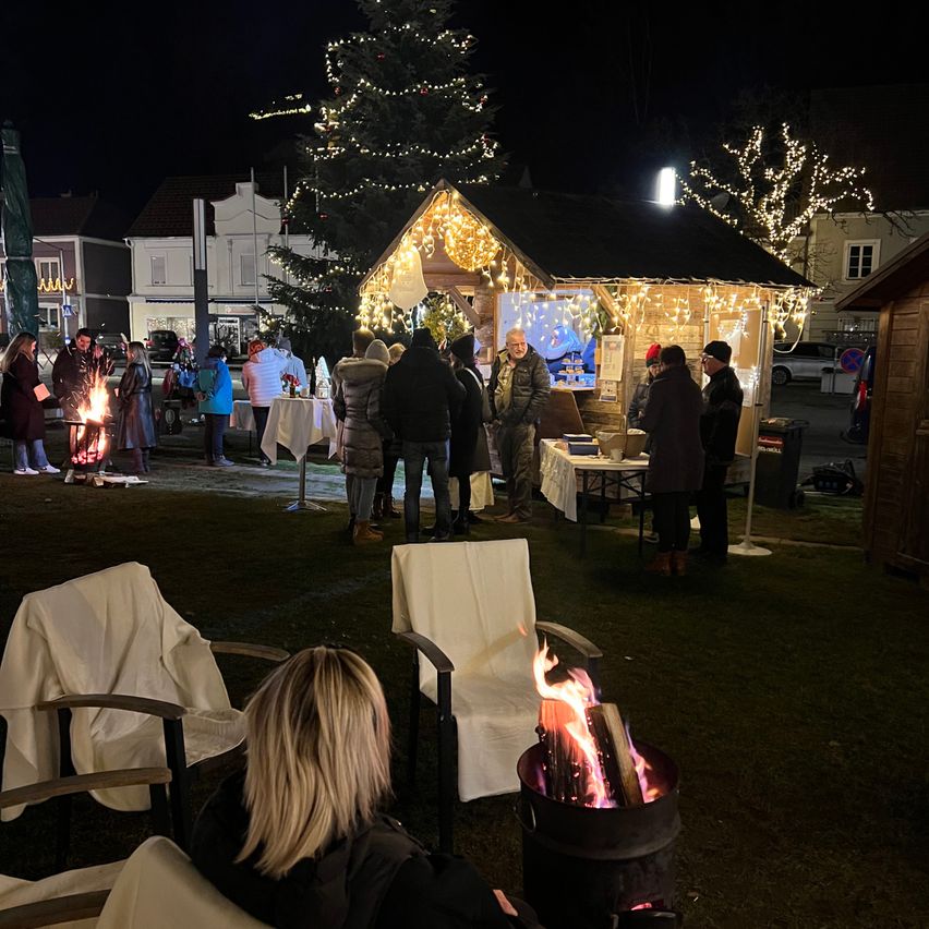 Eine Veranstaltung bei Nacht im Freien mit Menschen um ein Feuer, eine kleine Hütte mit Lichtern und einem Weihnachtsbaum.