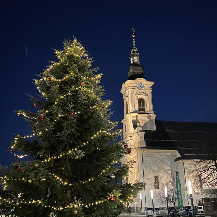 Ein großer Weihnachtsbaum ist mit Lichtern und Ornamenten verziert vor einer Kirche bei Nacht. Ein Glockenturm und eine Uhr sind sichtbar.
