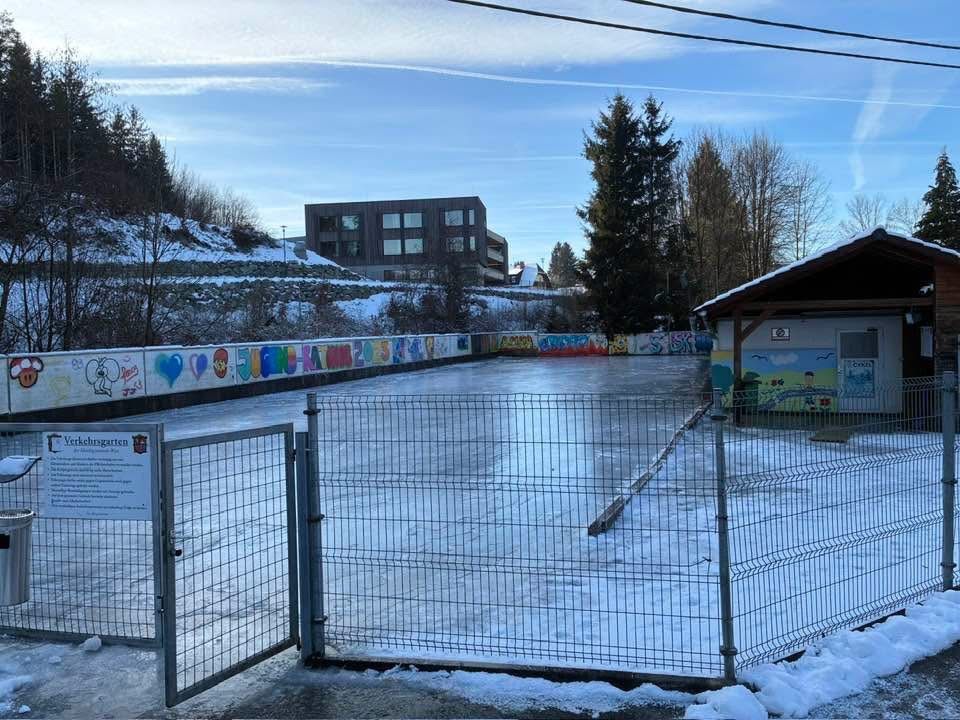 Eine leere, schneebedeckte Eisbahn mit farbenfrohen Graffiti an den umliegenden Mauern und einem Gebäude im Hintergrund unter einem blauen Himmel.