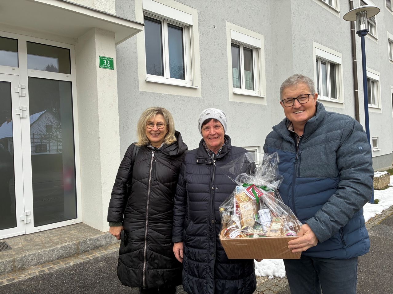 Drei Menschen stehen lächelnd vor einem Gebäude. Eine Frau links, eine andere in der Mitte und ein Mann rechts, der eine Schachtel mit einem Korb voller Lebensmittel hält.