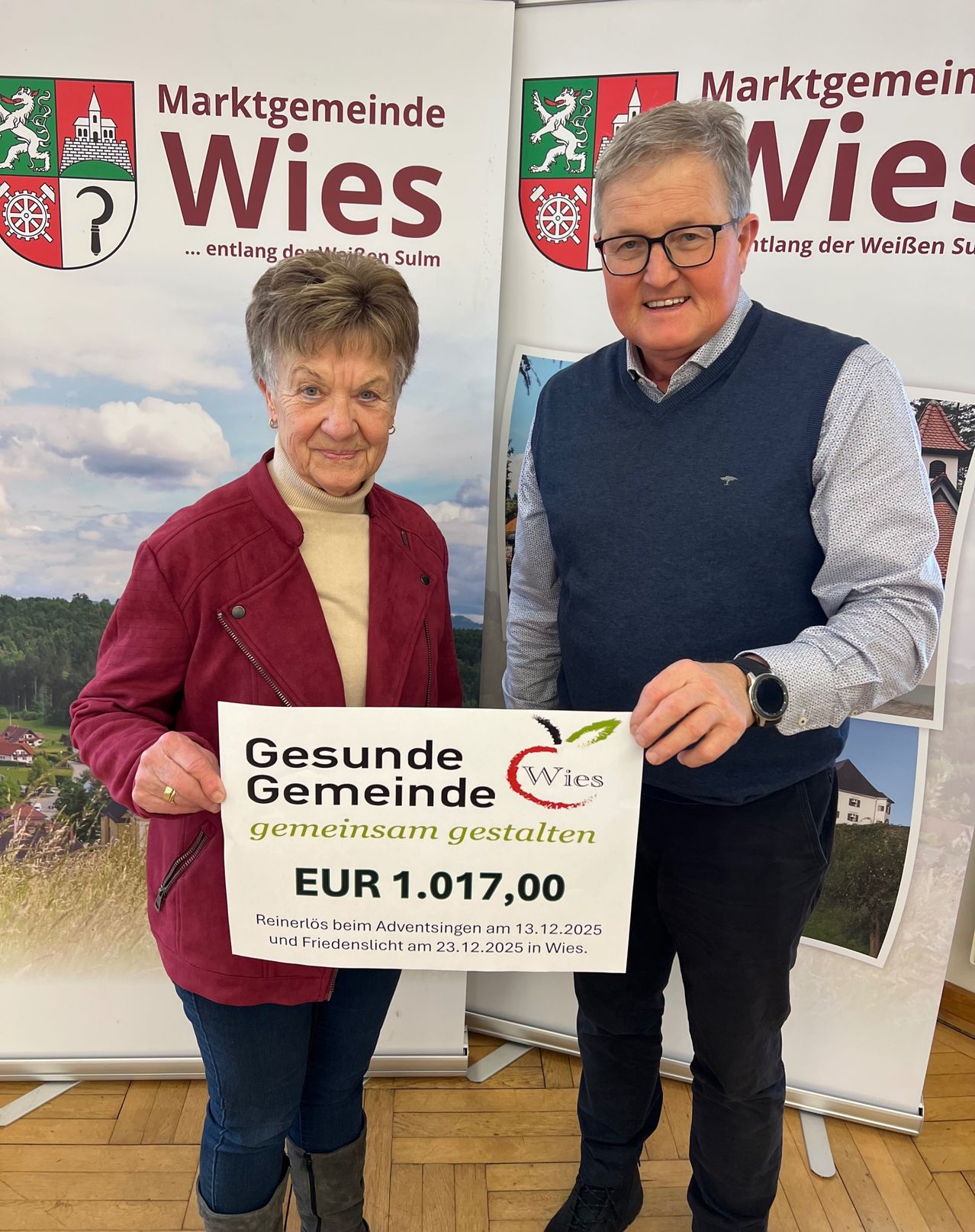 Eine ältere Frau hält ein Schild vor einem lächelnden Mann. Das Schild zeigt 'Gesunde Gemeinde gemeinsam gestalten EUR 1.017,00.' Sie stehen vor einem Hintergrund mit Bannern.