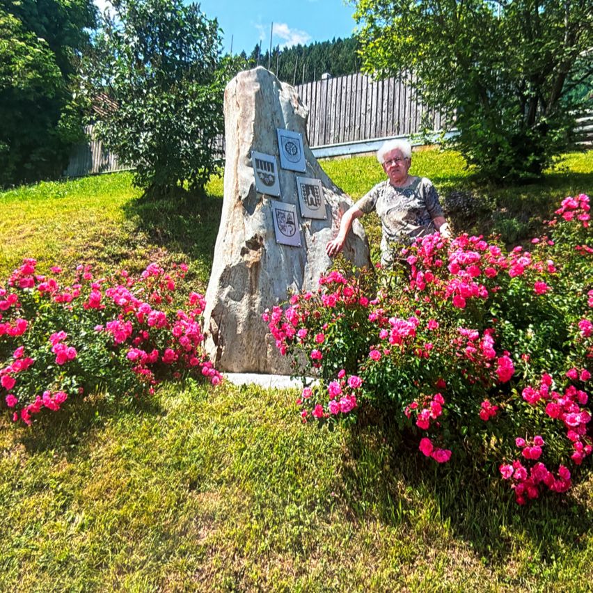 Eine ältere Frau steht neben einem großen Stein mit Tafeln, umgeben von leuchtend rosa Blumen. Der Hintergrund zeigt einen Holzzaun und üppiges Grün.