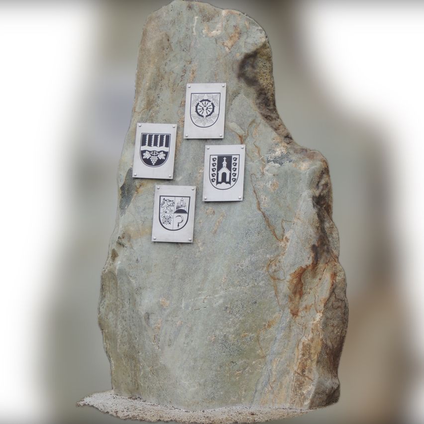 Ein großer grauer Stein mit vier auf seiner Oberfläche befestigten Emblemen. Die Embleme sind metallisch und rechteckig, jedes mit einem einzigartigen Design.