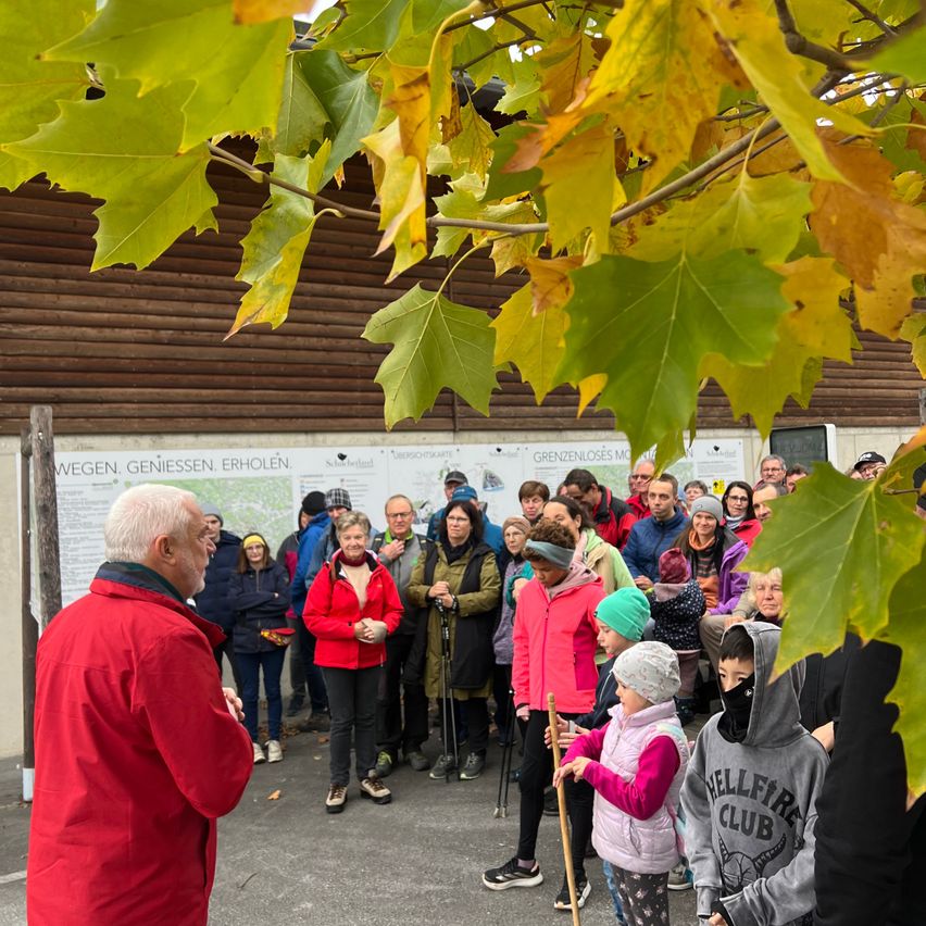 Eine Gruppe von Menschen ist unter einem Baum versammelt, möglicherweise für eine Versammlung. Der Mann im roten Mantel scheint zu sprechen. Viele von ihnen tragen Jacken, Mützen und Turnschuhe. Hinter ihnen steht ein Gebäude mit einem Plakat.