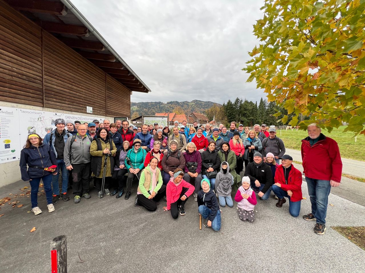 Eine Gruppe von Menschen posiert für ein Foto vor einem Holzgebäude. Einige stehen, andere sitzen. Sie sind in verschiedenen Winterkleidungen gekleidet, manche tragen Mützen und Stiefel. Im Hintergrund befinden sich Bäume und Berge unter einem bewölkten Himmel.