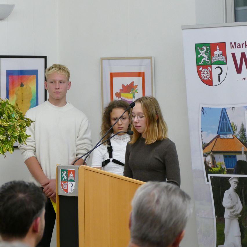 Zwei junge Menschen stehen an einem Podium mit Mikrofon. Einer trägt einen weißen Pullover und der andere ein schwarzes Langarmshirt. Dahinter hängt ein Banner mit einem Logo.