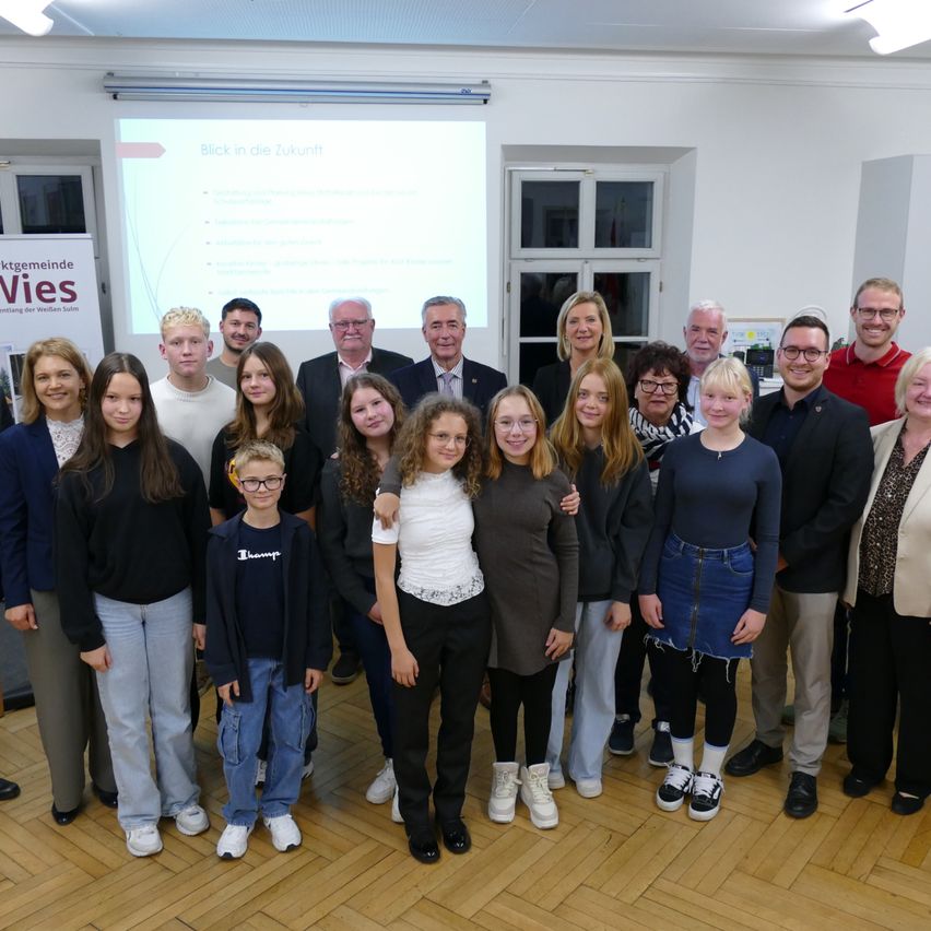 Eine Gruppe von Erwachsenen und Kindern posiert für ein Foto in einem Raum mit einer Projektionsleinwand, die eine Präsentation über die Zukunft zeigt.