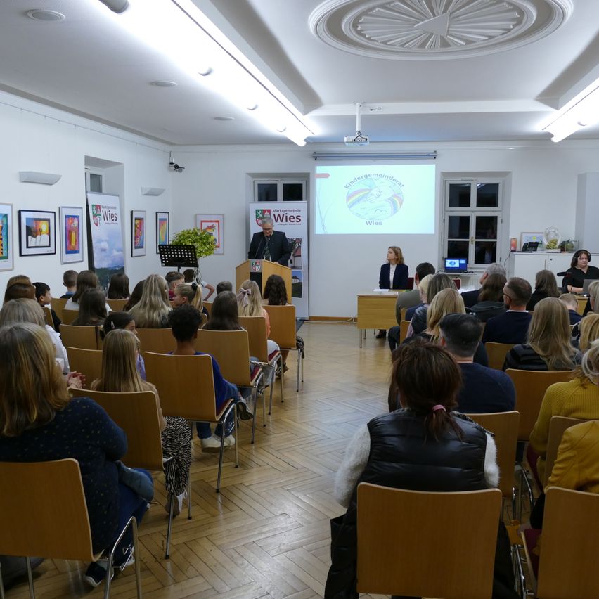 Ein Raum mit weißen Wänden und einer Projektionsleinwand zeigt eine Präsentation mit einem sprechenden Mann. Menschen sitzen in Stühlen, einige mit Kindern. Es gibt Bilder an der Wand, eine Pflanze und Tische mit Laptops.
