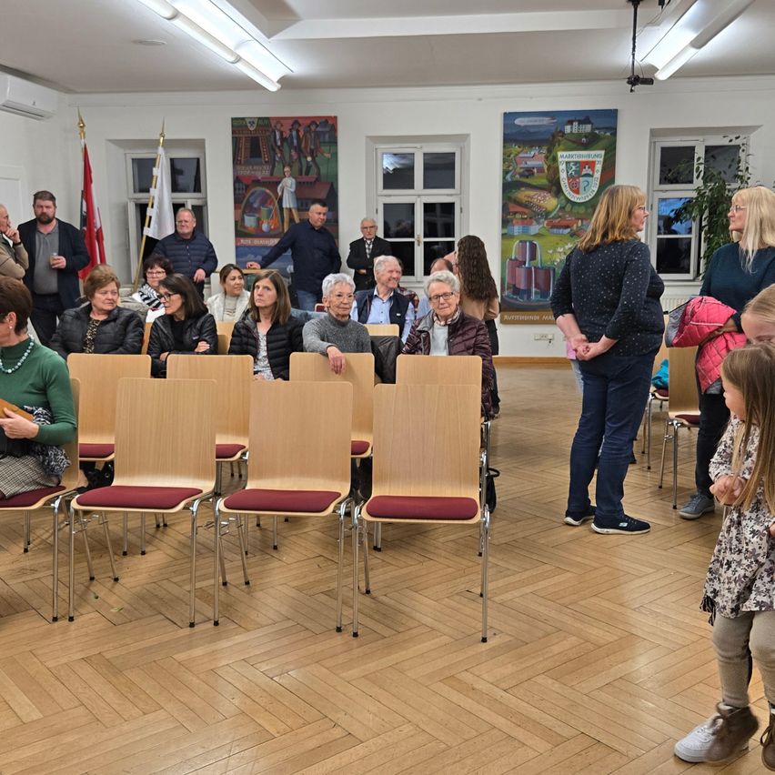 Eine Gruppe von Menschen sitzt in Stühlen in einem Saal. Einige stehen, andere sitzen. Zwei Frauen stehen hinter den Stühlen. Eine Frau hält eine Tasche. Zwei Banner hängen an der Wand.