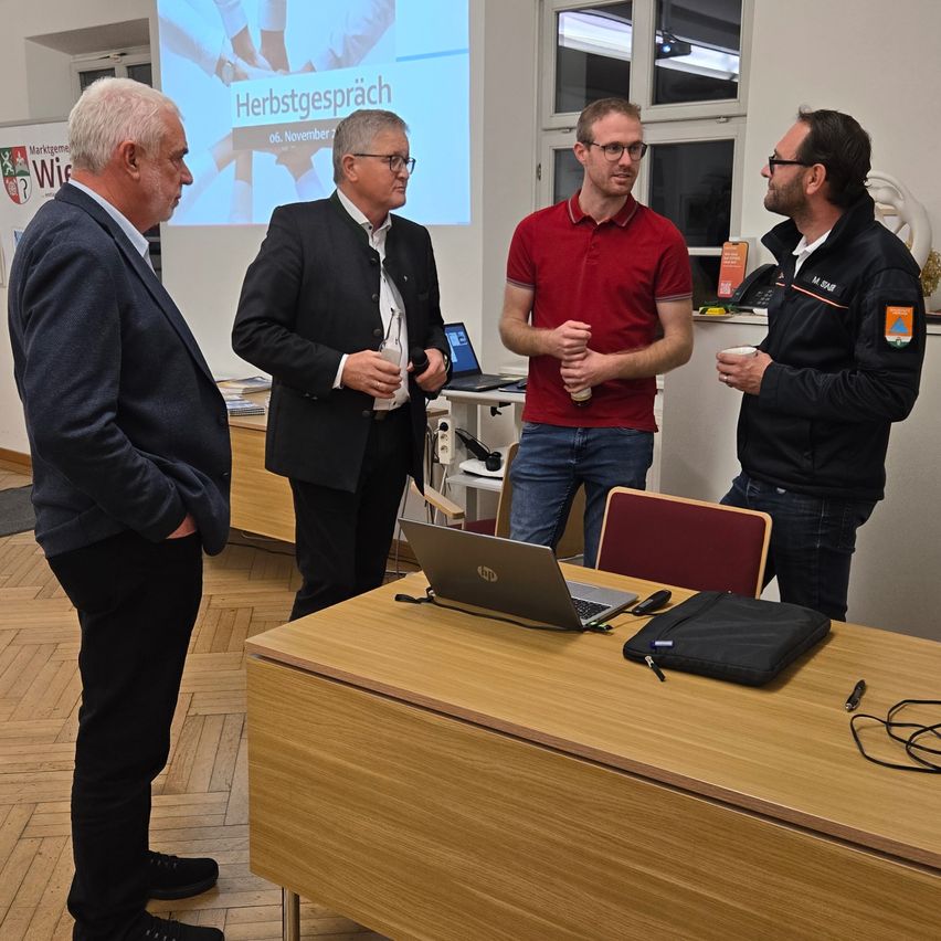 Vier Männer stehen in einem Büroraum. Eine Leinwand hinter ihnen zeigt den Text Herbstgespräch. Ein Laptop und eine Tasse stehen auf dem Schreibtisch.