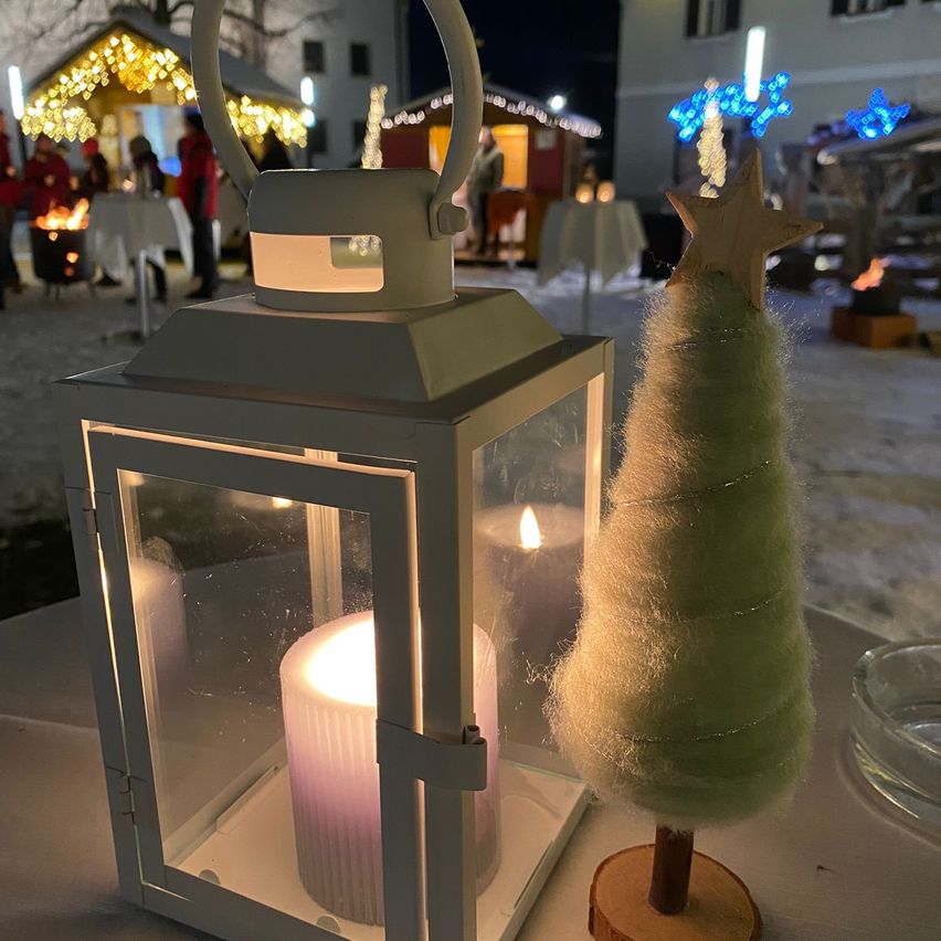 Ein Laternen mit einer brennenden Kerze steht neben einem kleinen, handgefertigten grünen Baum auf einem verschneiten Tisch, mit festlichen Lichtern im Hintergrund.