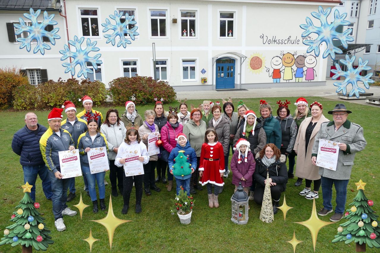 Eine Gruppe von Erwachsenen und Kindern in Weihnachtskleidung posiert für ein Foto vor einem Schulgebäude mit Schneeflocken und Kinderzeichnungen an der Wand. Einige halten Urkunden und Dekorationen in den Händen.