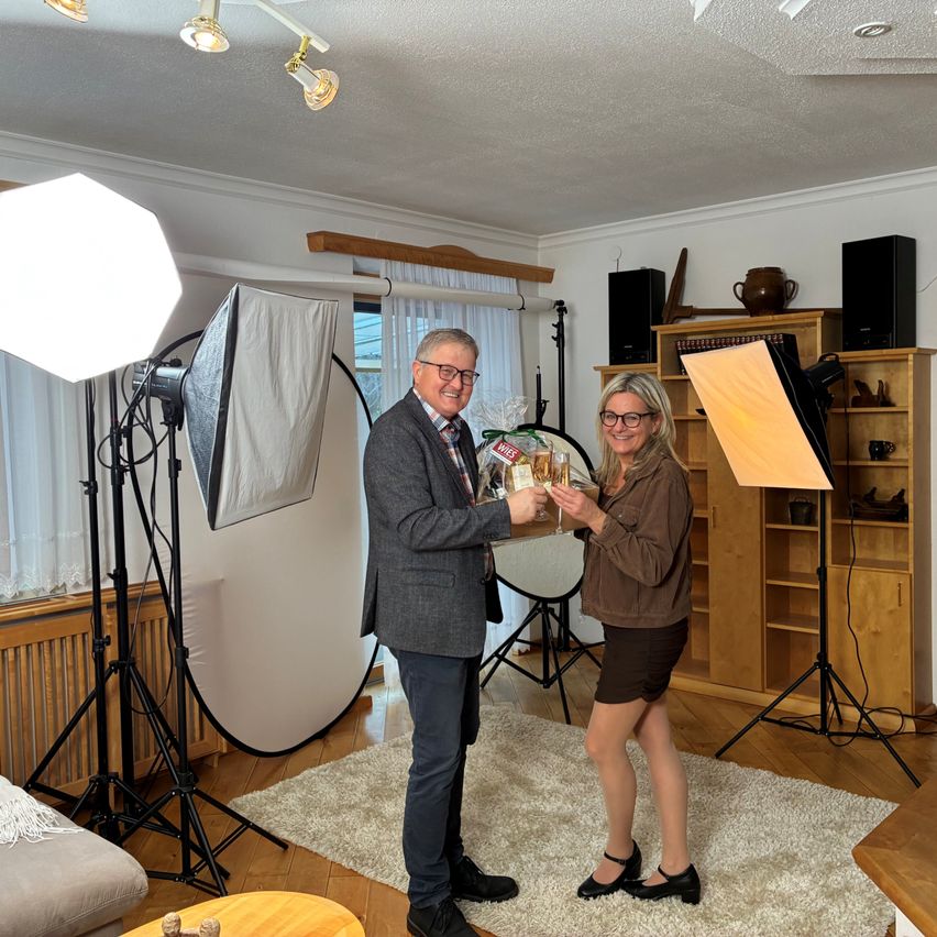 A man and a woman are holding a gift in a photo studio. The man wears a gray suit and glasses, and the woman wears a brown shirt and glasses. They are surrounded by photography equipment.