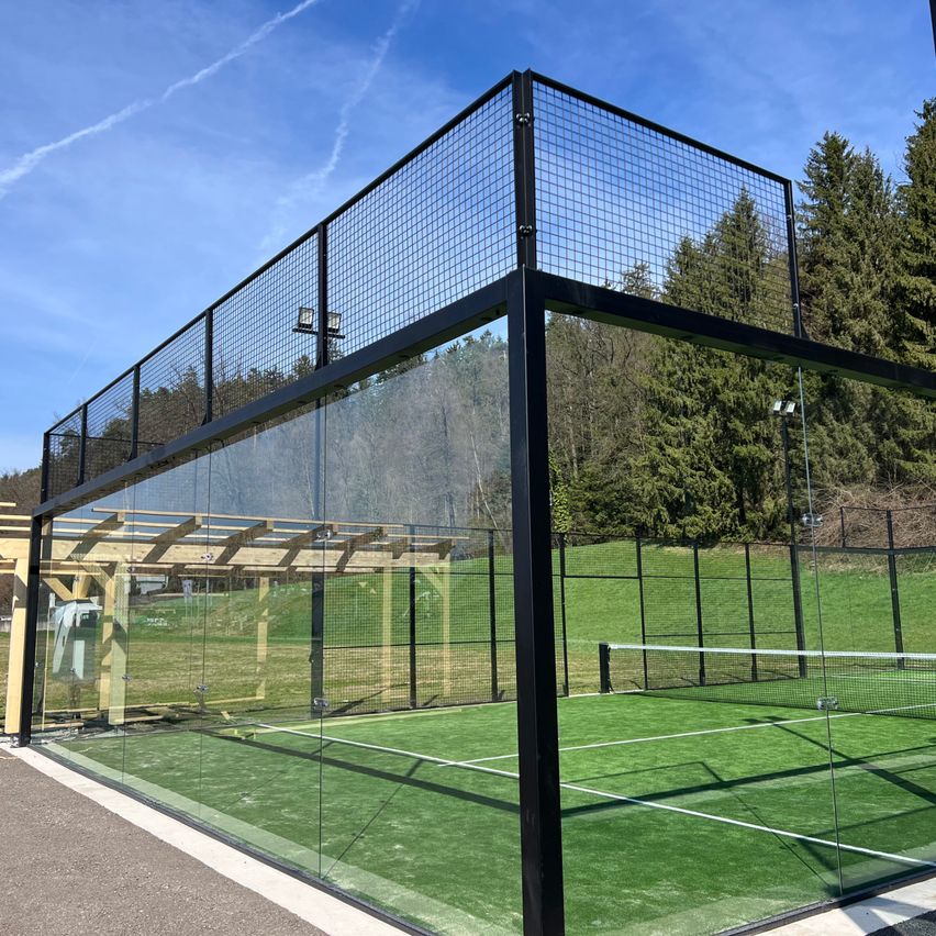 An enclosed tennis court with glass walls, a net, and a shed, surrounded by green grass and trees under a clear blue sky.