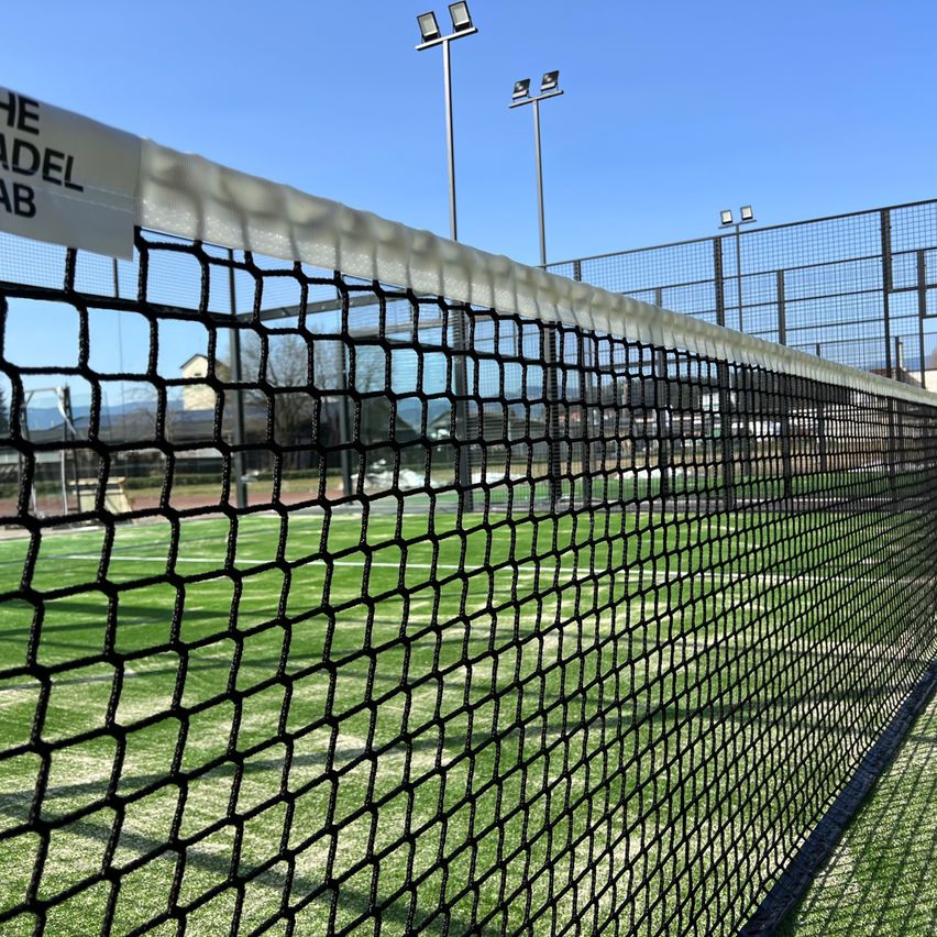 A tennis court with a net and grass field, featuring a label reading 'THE ADE' and surrounded by streetlights.