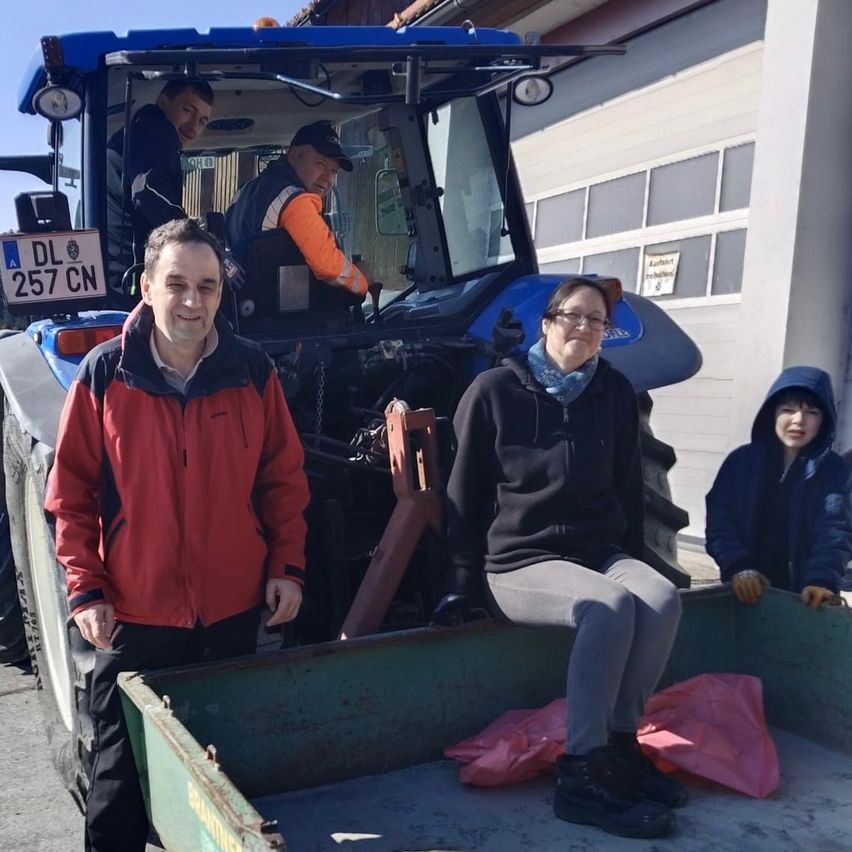 A group of people stand in front of a tractor. A man in red is on the left, a woman in black is sitting in the back, and a child is on the right.