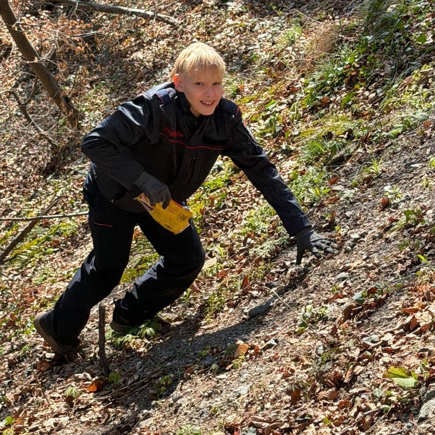 A boy in a black jacket is climbing up a steep, leafy hill with a yellow object in hand.