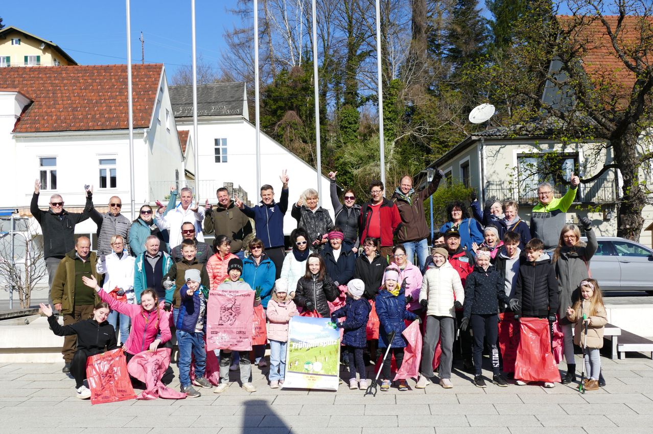 A large group of people poses for a photo outside. Many are waving. Some hold pink bags, one holds a pink banner. Trees and buildings are in the background.