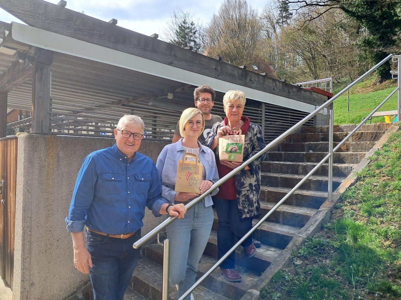 Four people stand on stairs outdoors with bags. A man in a blue shirt and a woman in blue jeans hold hands. Behind them, two others, a man in a maroon shirt and a woman in a floral coat, hold bags.