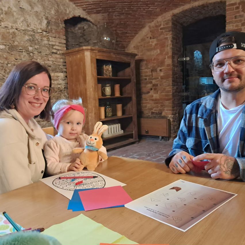 A family of three with a young girl holding a stuffed toy sitting at a table with coloring sheets and pencils on it. Behind them, a cabinet and a brick wall are visible.