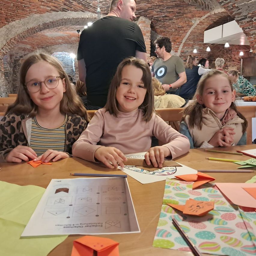 Three young girls sit at a table, smiling and engaged in crafting activities. They have a variety of colored papers and pencils in front of them. In the background, other people are seated and standing.